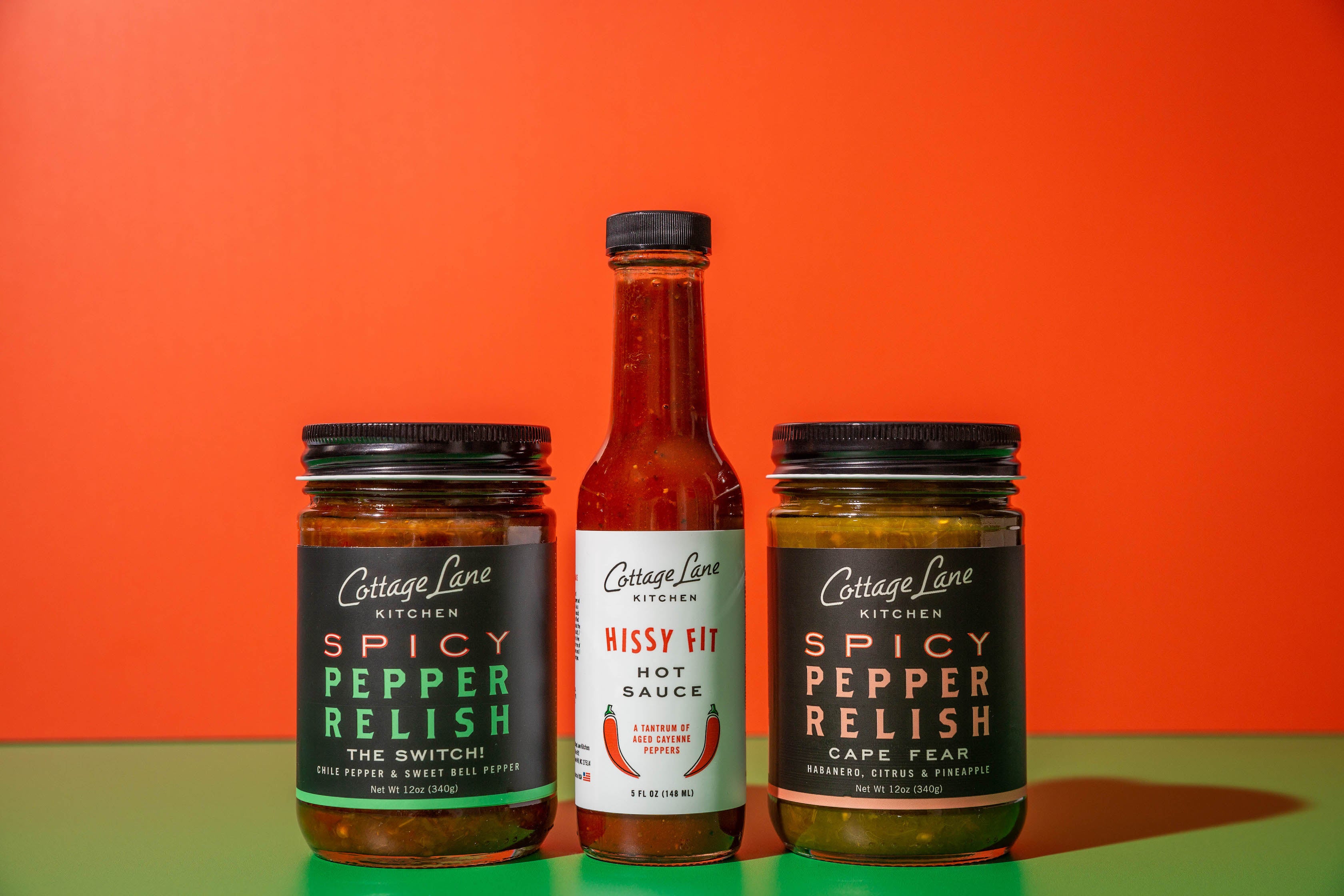 A photo of Cottage Lane Kitchen Spicy Pepper Relishes, Cape Fear and Get Me A Switch, and Hissy Fit Hot Sauce on green shelf with bright red background