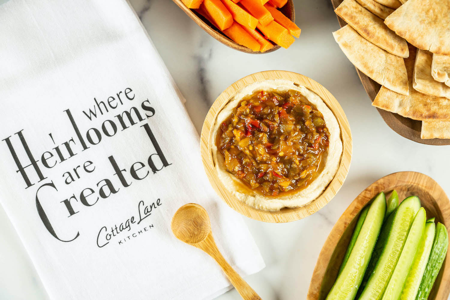 Spicy Pepper Relish on Hummus with carrots cuccumbers and pita bread and a dish towel that says where heirlooms are created Cottage Lane Kitchen